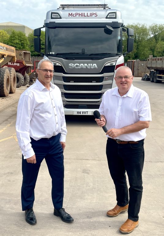 Two people stood in front of a parked HGV, one is holding a microphone.