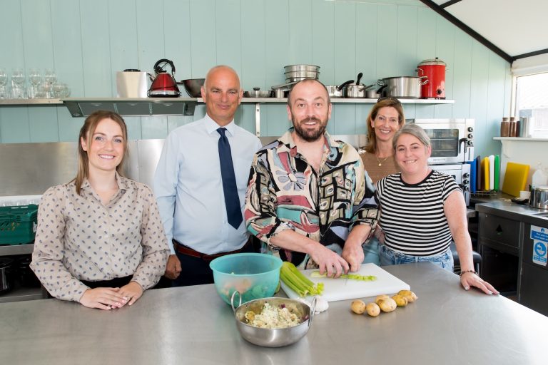Five people stood in a kitchen smiling. The person in the middle is cutting up vegetables and putting them in a bowl.