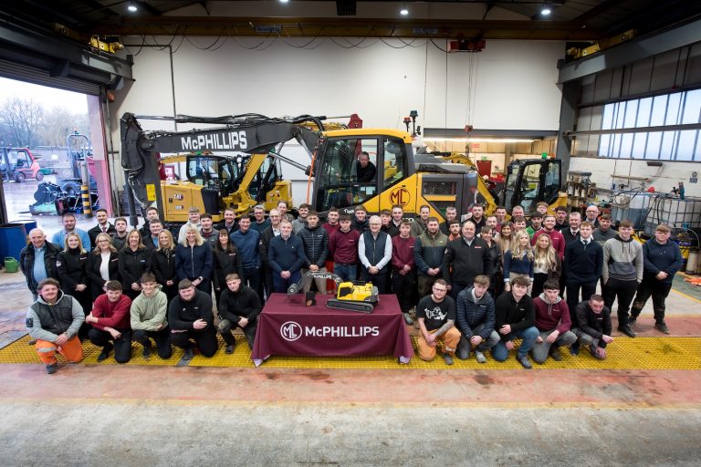 A large group of people stood in a warehouse in front of a digger. In front of them is a table with a small replica digger on it.