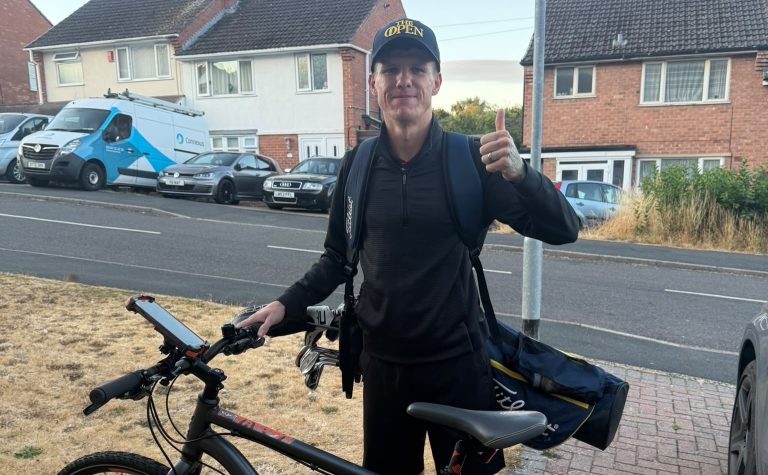 A person stood with a bicycle and a bag of golf clubs over their shoulder.