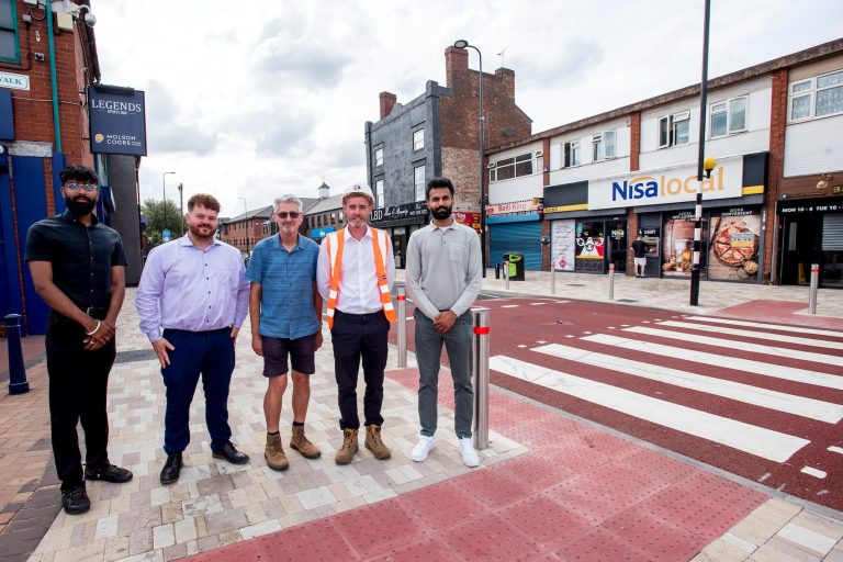 Four people stood together in a line opposite a row of high street shops.